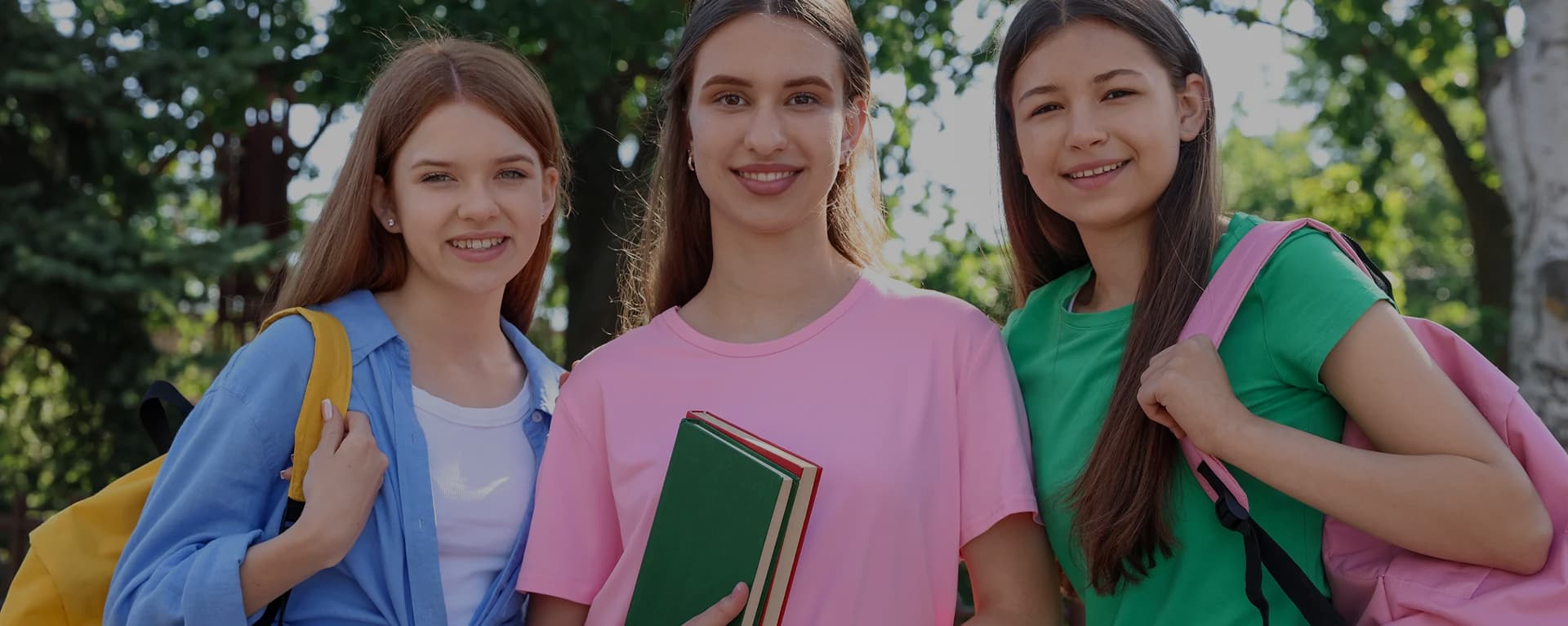 Three smiling teenage girls outdoors with backpacks and books